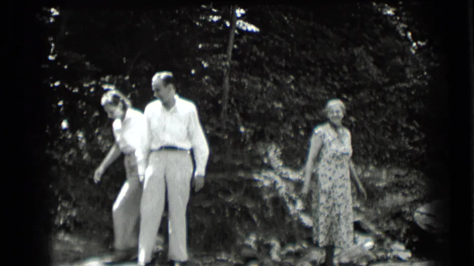 1939 Clear Lake frame with three adults in casual clothing moving through a wooded natural area near a small water edge.