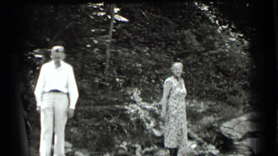 At Clear Lake, California in 1939, a man and woman stand apart on rocks near a wooded streambank.