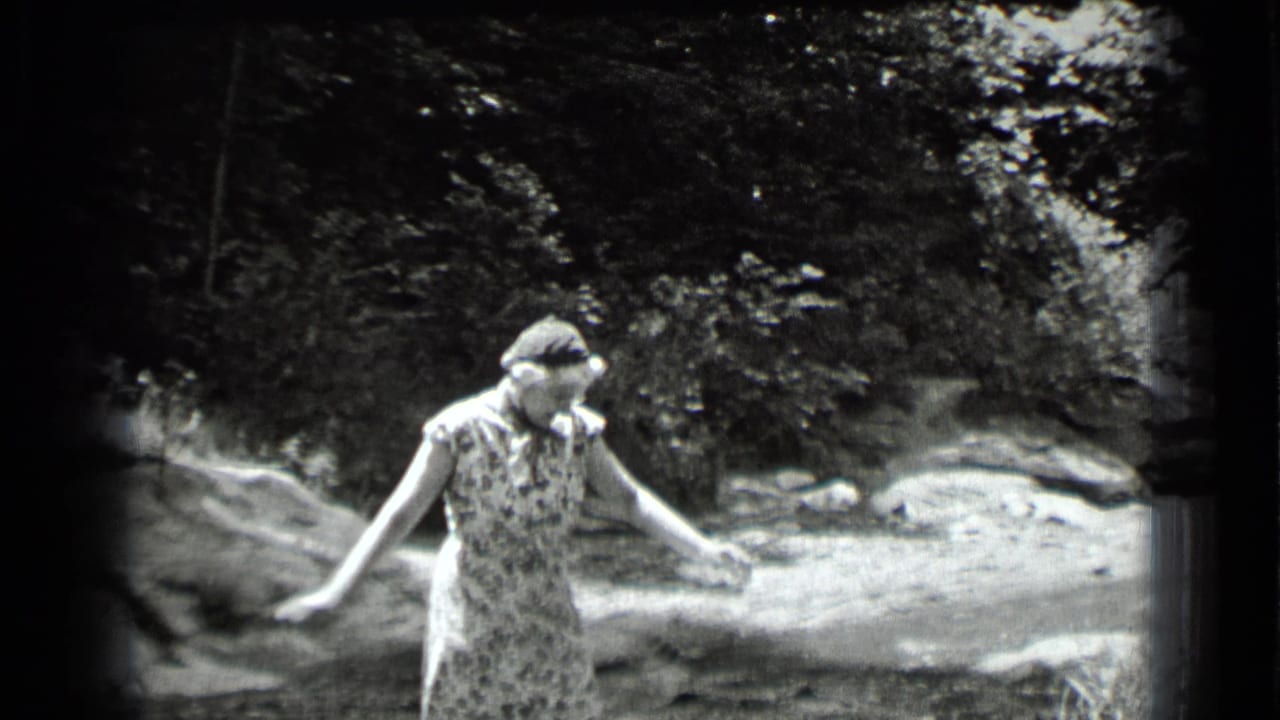 Woman in a patterned dress balances near rocks beside a stream.