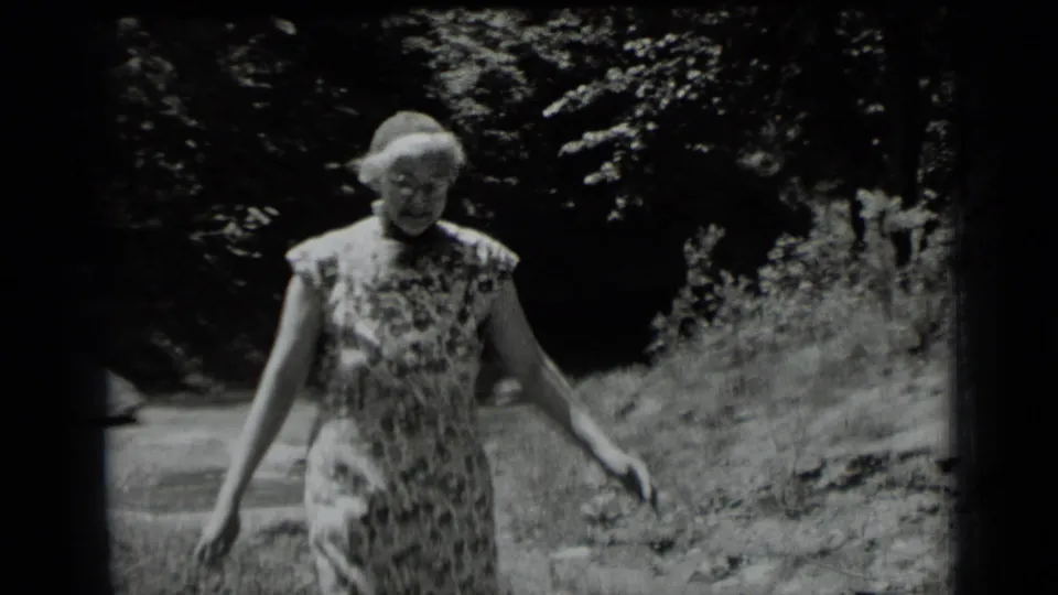 Adult woman in a floral dress walks through a shaded Clear Lake, California forest in 1939 home-movie footage.