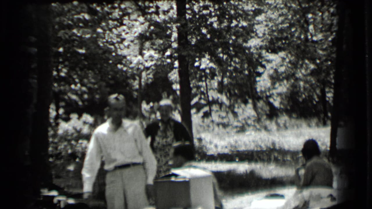 Adults gathered around outdoor tables in a wooded park, with one man walking in the foreground.