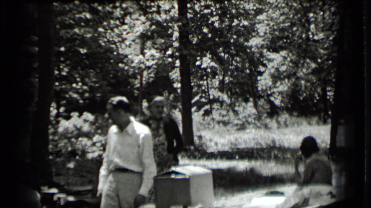 A man in a light shirt stands near a table while other adults appear behind him among trees.