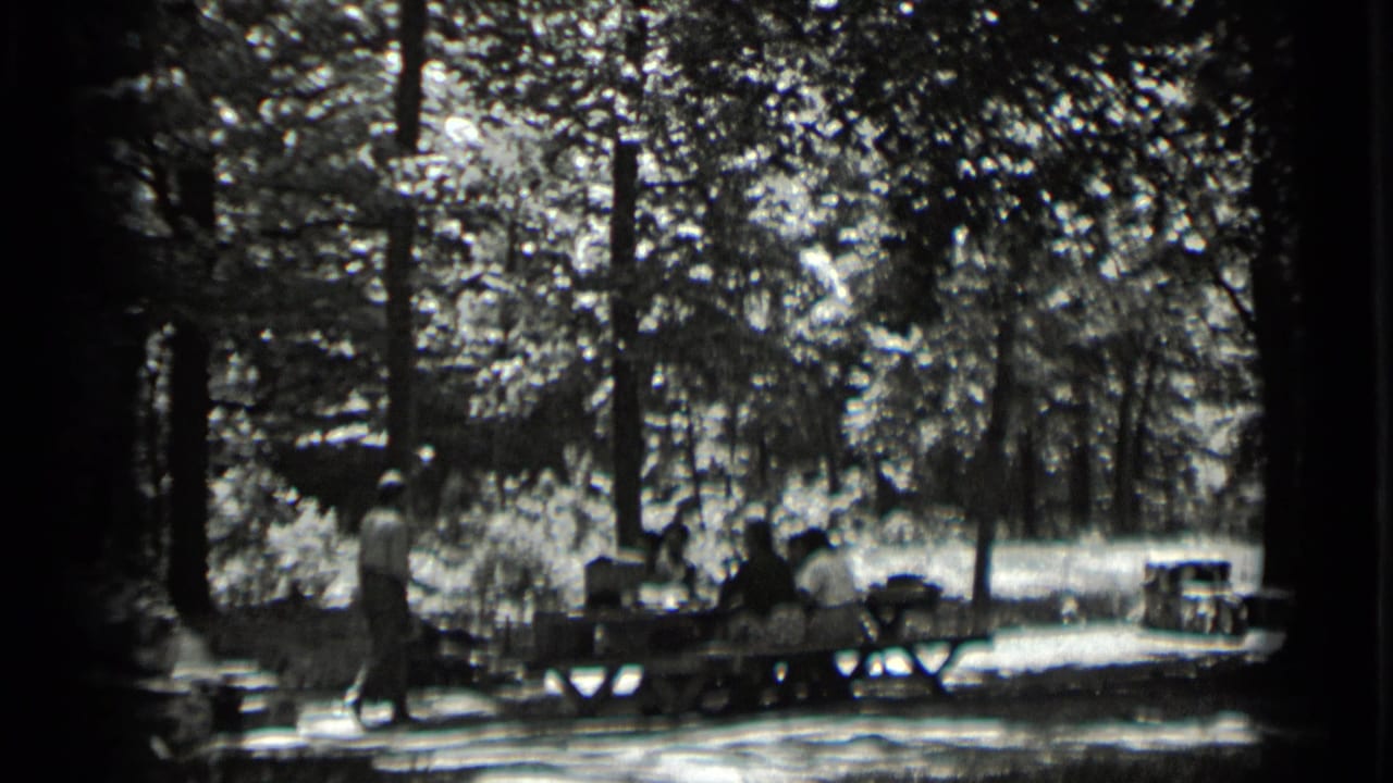 1939 Clear Lake frame with several people at a picnic table under trees and a standing figure nearby.