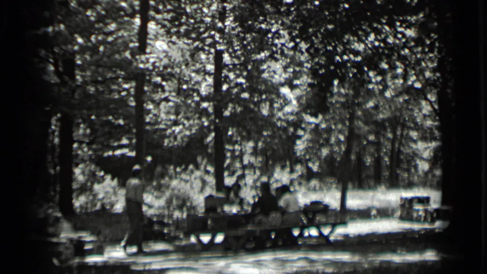 1939 Clear Lake frame with several people at a picnic table under trees and a standing figure nearby.