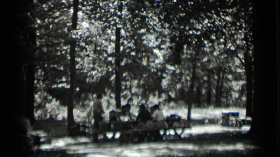 1939 Clear Lake home-movie frame showing a shaded picnic table, seated figures, and belongings under tall trees.