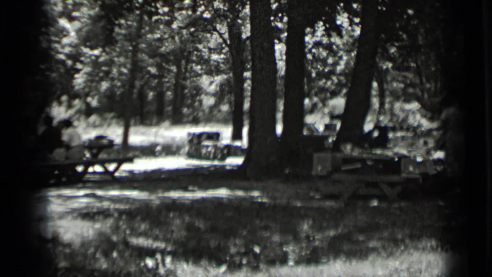 Large tree trunks frame a Clear Lake campsite where people sit near picnic tables in black-and-white 8mm footage.
