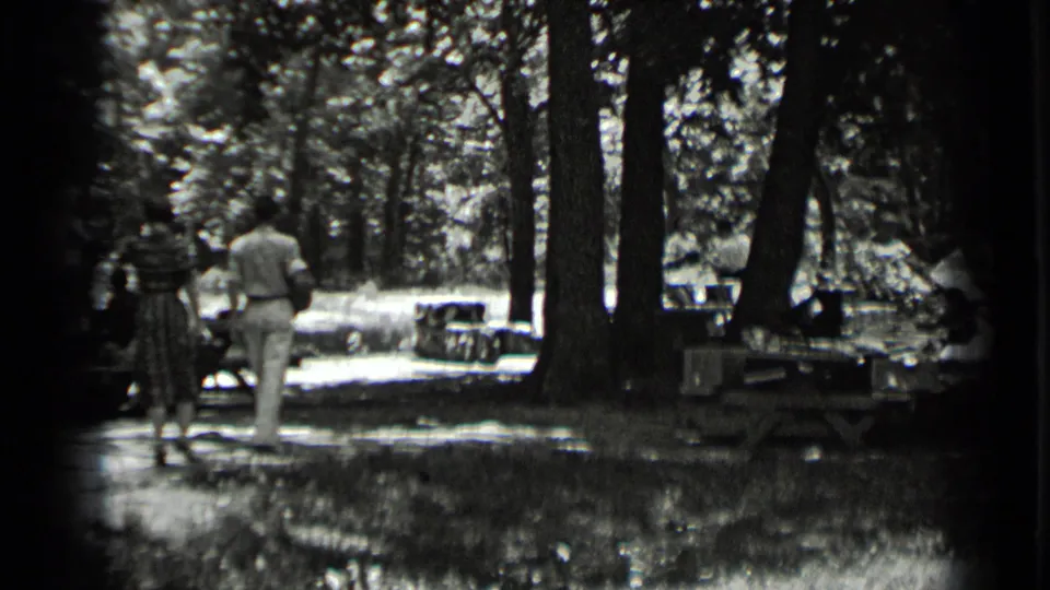 A man in light clothing and a woman in a patterned dress walk through the Clear Lake wooded picnic area in 1939 footage.