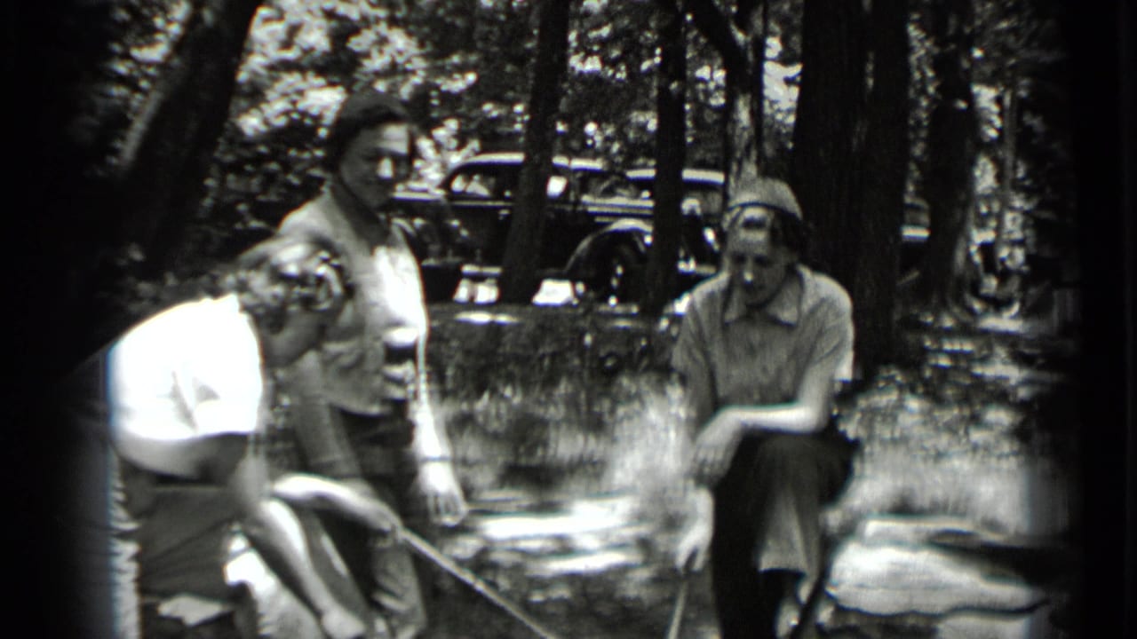 Three women stand and crouch near a campfire in a wooded clearing.