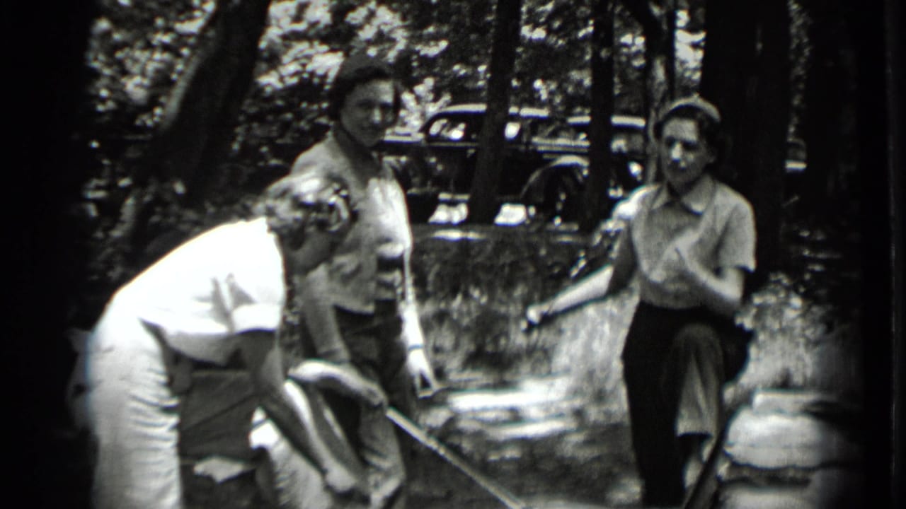 Women lean toward a small fire while holding long sticks outdoors.
