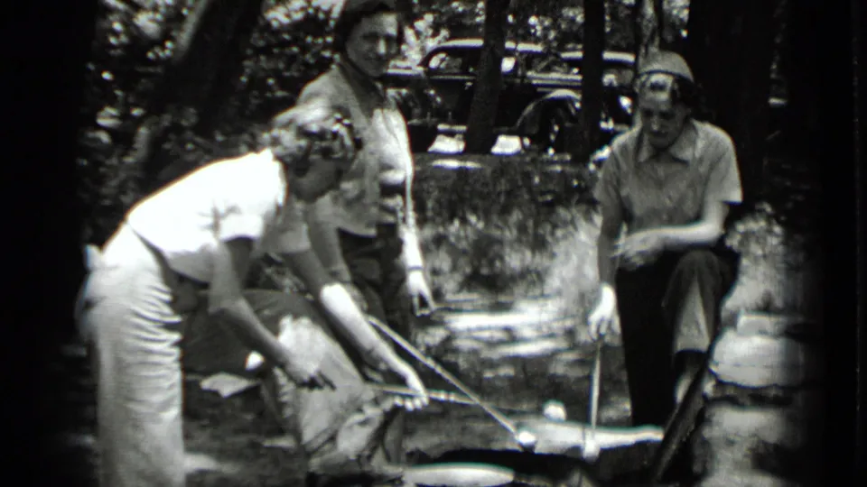 A woman crouches near the campfire while two others stand or sit close by during a 1939 Clear Lake outdoor gathering.