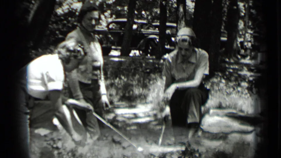 The women continue roasting over the fire in a wooded Clear Lake camp scene, with rocks and a vintage car in the background.