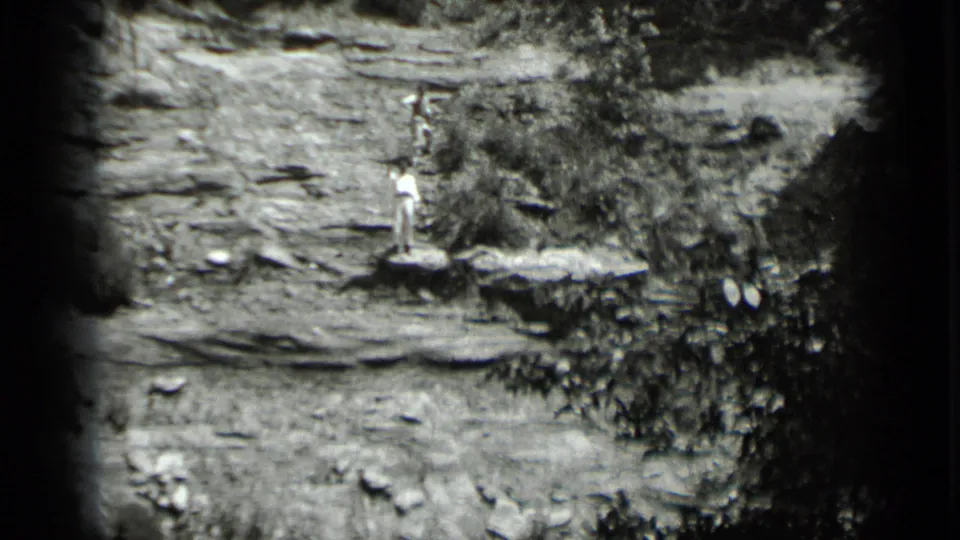 Black-and-white 8mm frame from Clear Lake shows a person standing on a cliffside rock ledge with vegetation to the side.