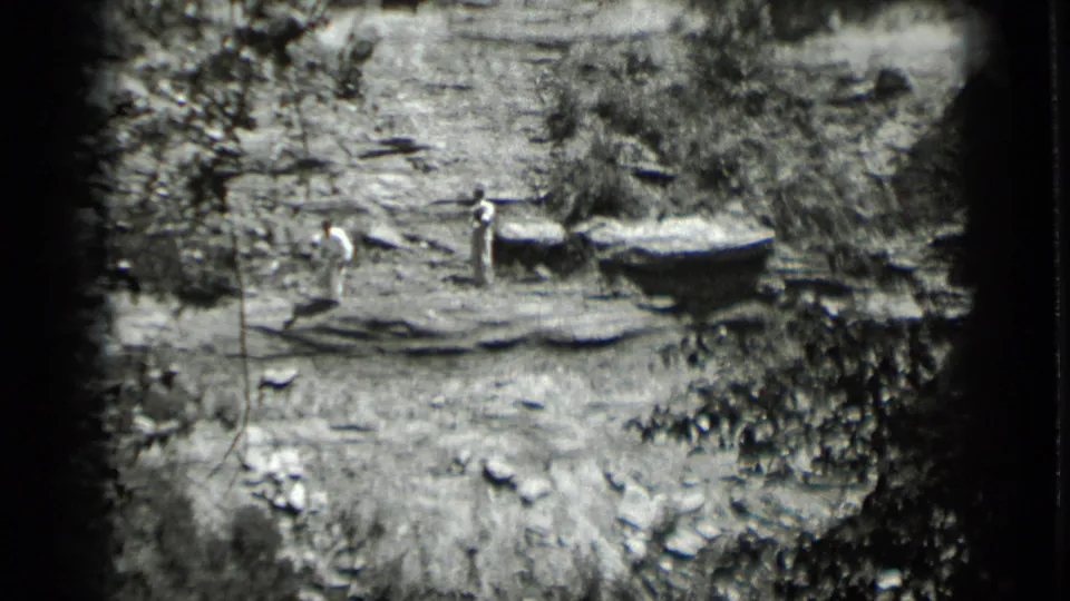 1939 Clear Lake home-movie frame with two men near rough wooden platforms beside brush, rocks, and wet shoreline terrain.