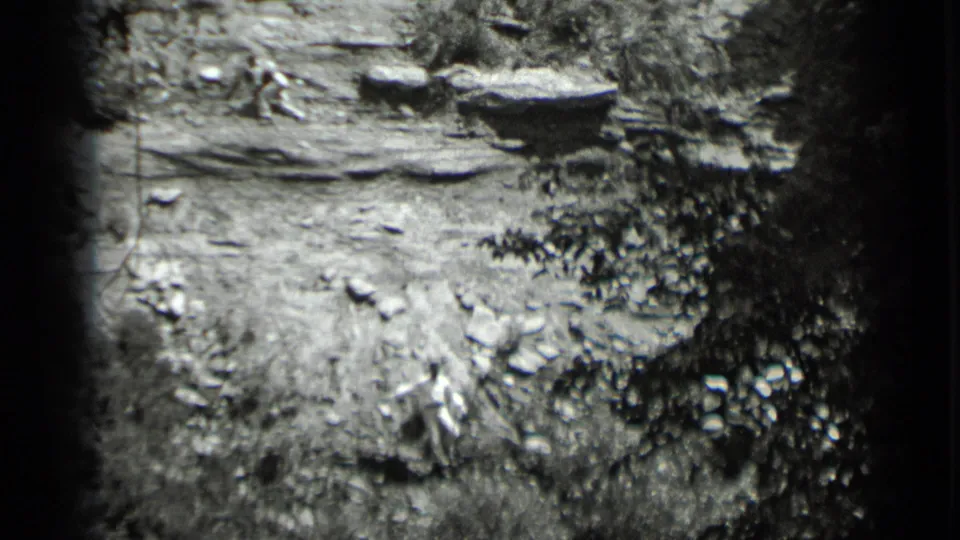 Black-and-white Clear Lake frame with a figure in light clothing crossing rocky terrain near dense foliage.