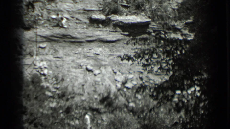 Archival 8mm frame of a steep, brush-lined rocky slope at Clear Lake with a small figure in light clothing partly visible.