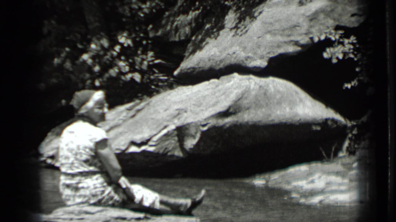 1939 Clear Lake home-movie frame showing a person in light clothing seated near shallow water below large rocks.