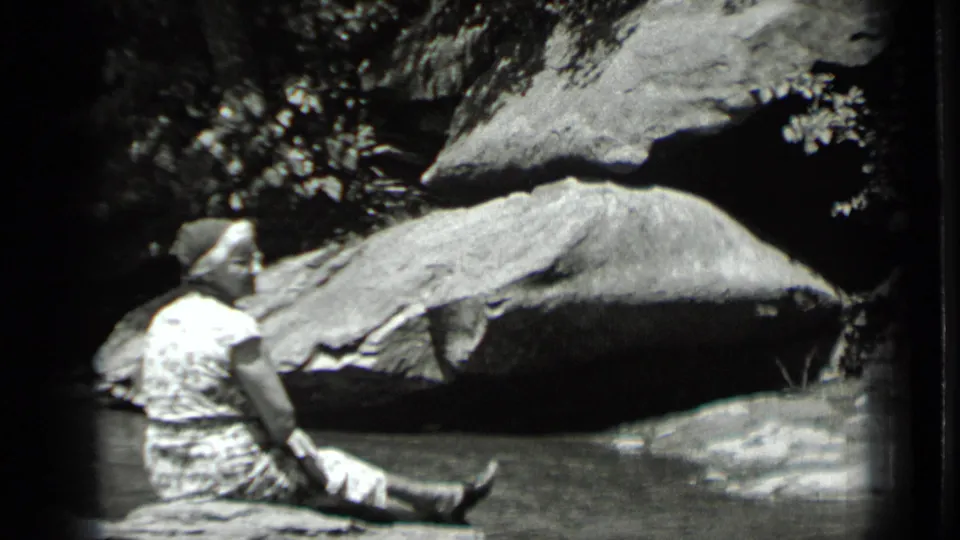 1939 Clear Lake home-movie frame showing a person in light clothing seated near shallow water below large rocks.