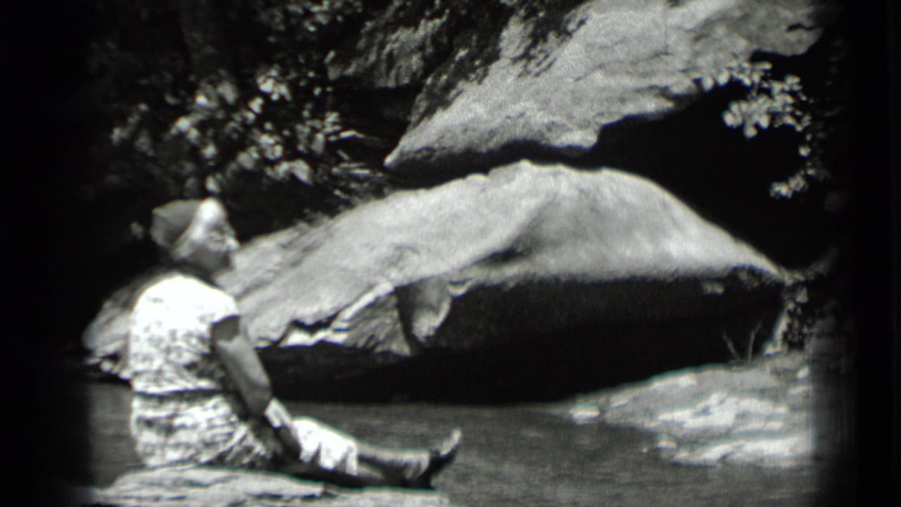 In 1939 Clear Lake, California footage, a woman sits beside shallow water under large rocks and shaded foliage.