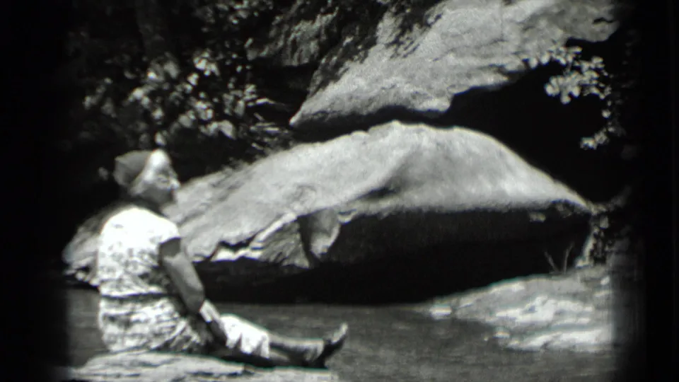 In 1939 Clear Lake, California footage, a woman sits beside shallow water under large rocks and shaded foliage.