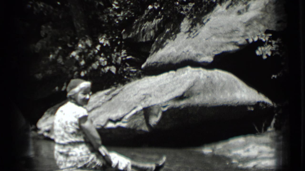 Seated woman turns near water with boulders and trees behind her.