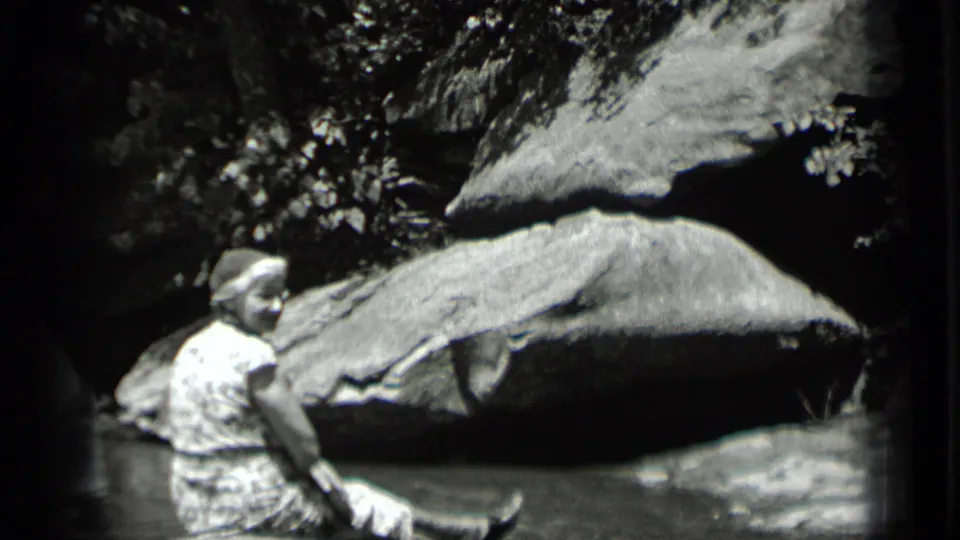 A seated woman turns toward the camera in 1939 Clear Lake footage, framed by shallow water, boulders, and trees.