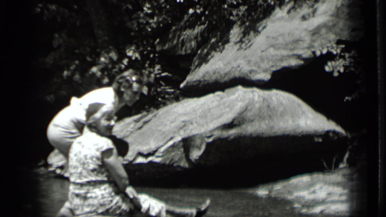Two adult women appear by the water in 1939 Clear Lake footage, one seated and one bending near her against large rocks.
