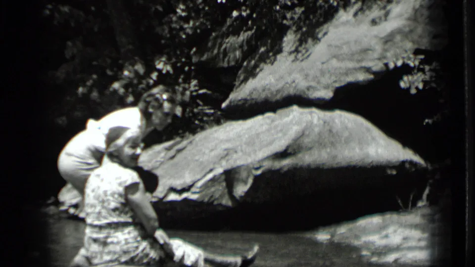 Two adult women appear by the water in 1939 Clear Lake footage, one seated and one bending near her against large rocks.