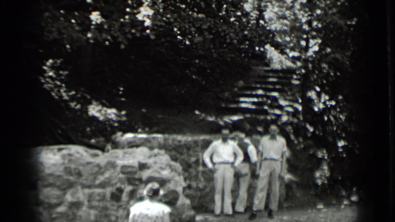 Two men stand near a stone wall while a seated figure faces them in a wooded ruin.