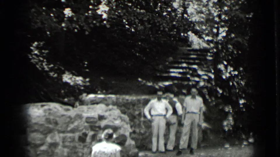 In 1939 Clear Lake, California footage, two men in light clothing stand by stonework as another person sits in the foreground near trees and steps.