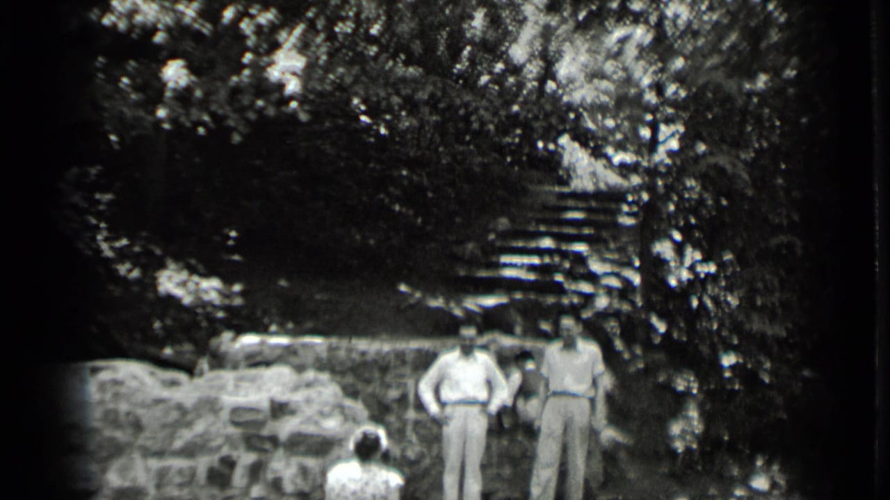 A 1939 Clear Lake home-movie frame shows men near a rustic stone wall, with a seated visitor in front and shaded steps behind them.