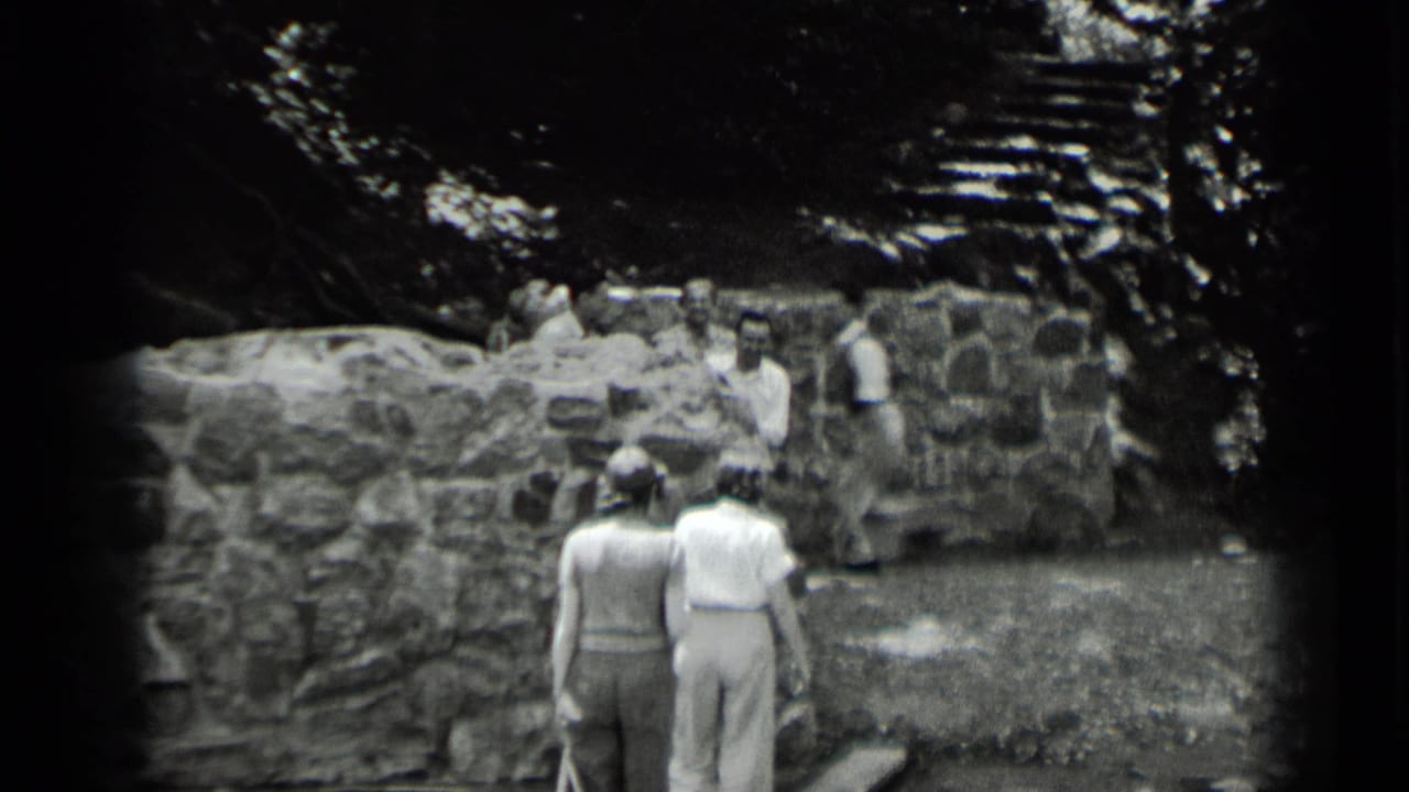 Two women, seen from behind, walk toward men beside a low stone wall and steps in 1939 Clear Lake, California footage.