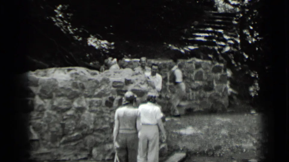 Two women, seen from behind, walk toward men beside a low stone wall and steps in 1939 Clear Lake, California footage.