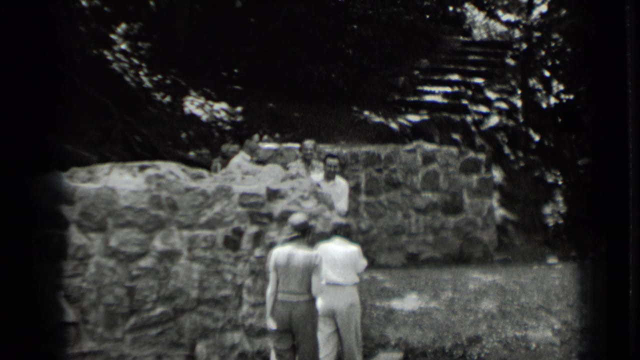 Two women walk toward people gathered beside a low stone wall under trees.