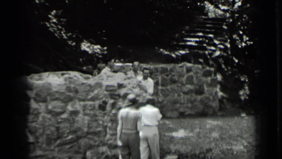 At Clear Lake, California in 1939, two women approach adults gathered near a rustic stone wall in a wooded area.