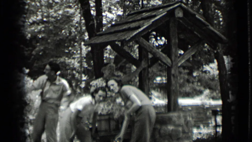 Young men and women in 1939 Clear Lake footage stand and bend near a rustic wooden well under trees.