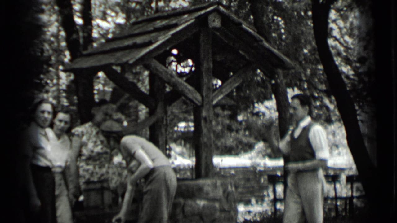 Adults stand near a rustic wooden shelter in a wooded area.