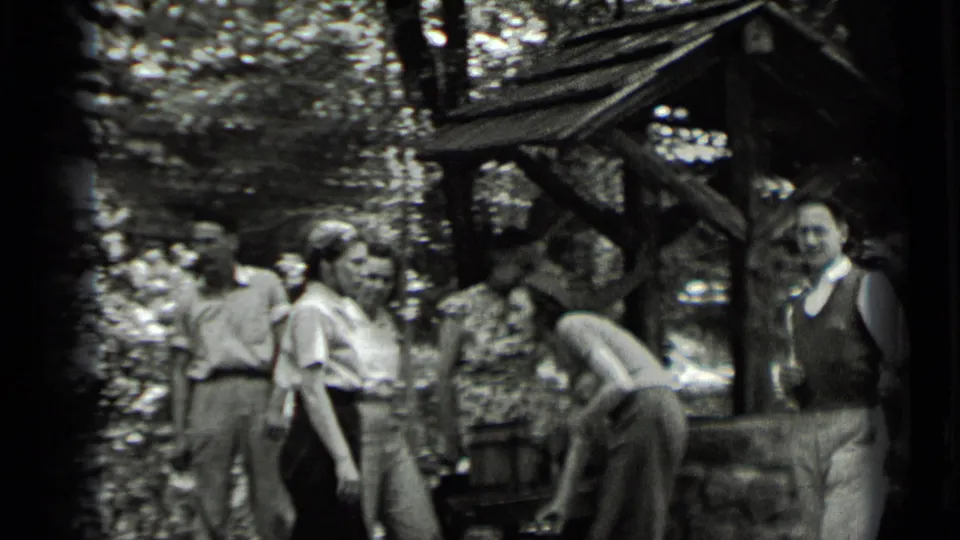 Adults in 1930s attire appear near a rustic shelter at Clear Lake, with one person bending and others facing the camera or each other.