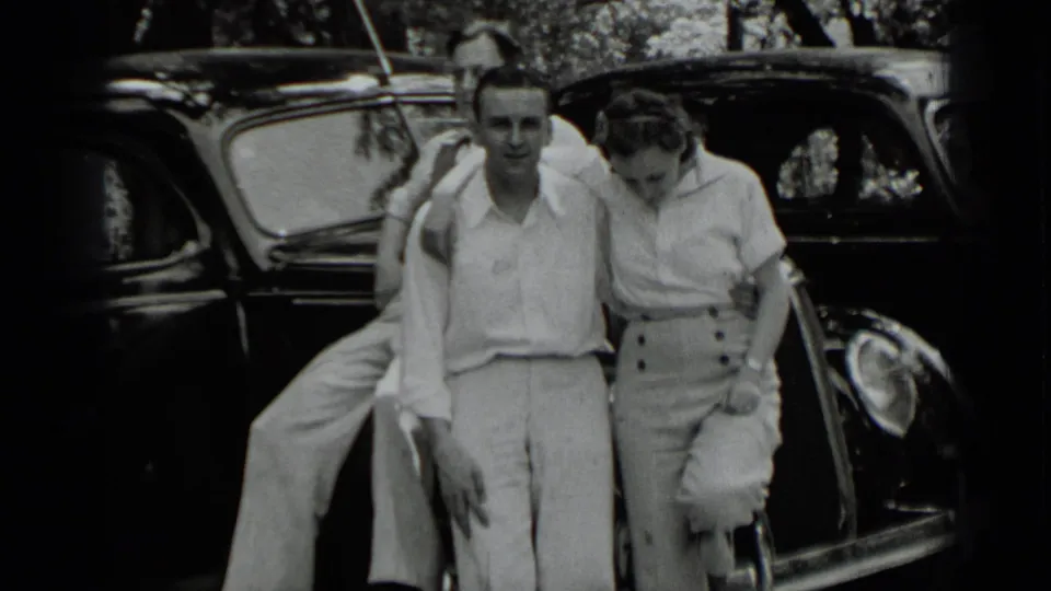 A man and woman in 1930s attire stand arm in arm beside a dark car during the Clear Lake home movie.