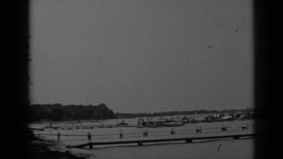1939 Clear Lake home-movie frame with dock slips and small boats spread across calm water near a wooded shoreline.