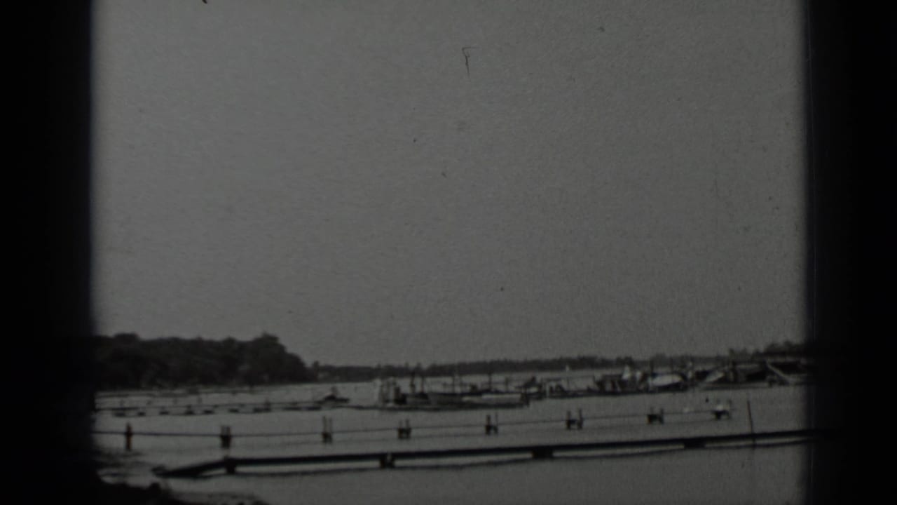 Black-and-white lake view with moored boats, a long pier, distant hills, and film borders.