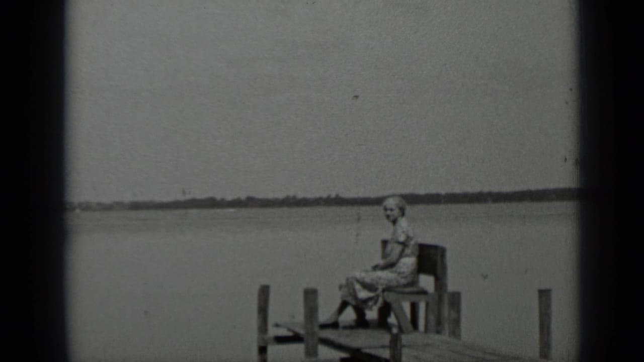 Older woman seated on a wooden dock beside a wide lake with a distant shoreline.
