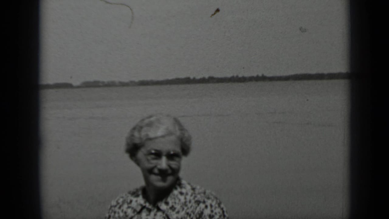 Older woman with glasses smiles beside a lake with distant shoreline behind her.