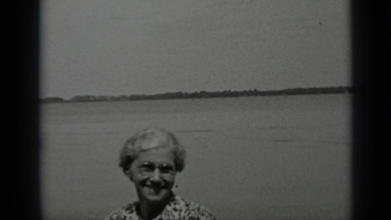 Older woman smiles near the lower frame with open water and shoreline behind her.