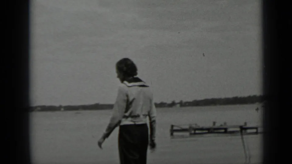 A woman in a light sweater and dark trousers walks beside Clear Lake, California, with water, distant shore, and a small dock visible.