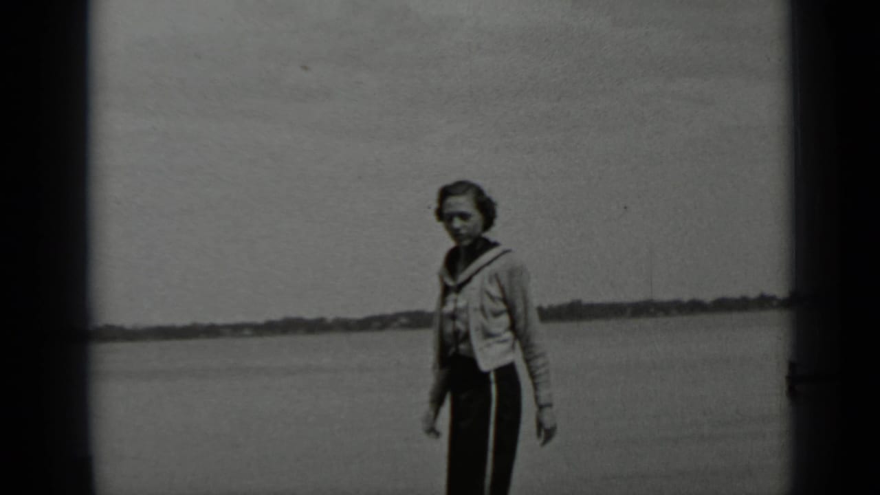 Woman near a lake turns toward the camera with open water behind her.