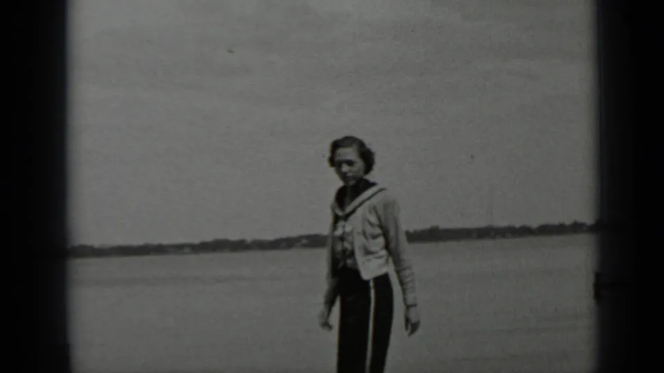 A woman near the Clear Lake shoreline faces the camera, wearing a light sweater over dark trousers with the lake stretching behind her.