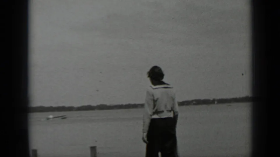 The woman is filmed from behind near Clear Lake, with broad water, a low shoreline, and a small object near the waterline in view.