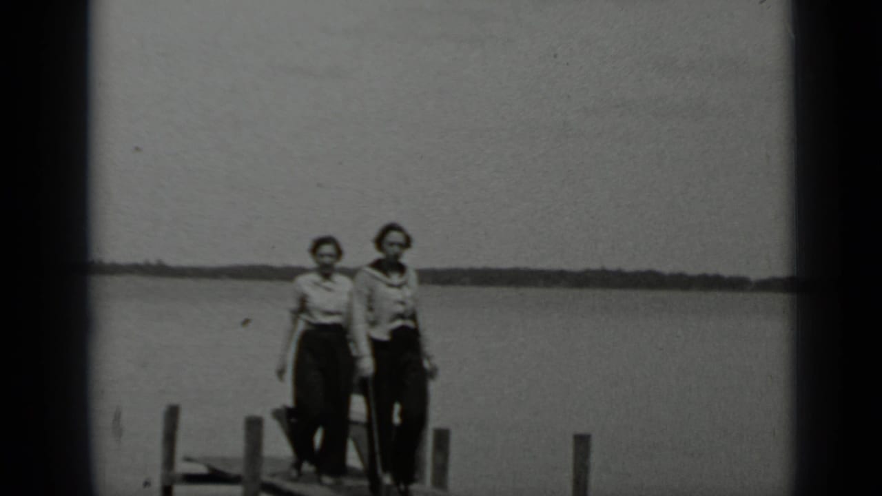 Two women stand or walk on a wooden pier with lake water behind them.