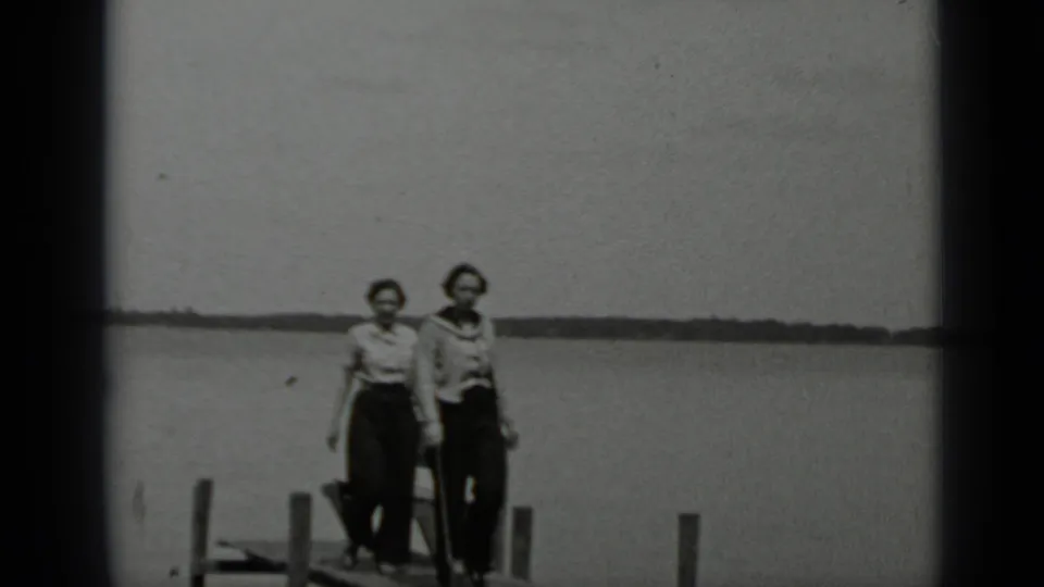 Two women on a Clear Lake wooden pier face the camera, with calm water, dock posts, and a distant shoreline visible behind them.