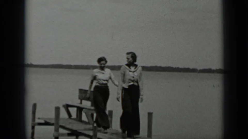 Two women in casual 1930s clothing walk side by side on a Clear Lake pier, passing dock furniture with open water behind them.